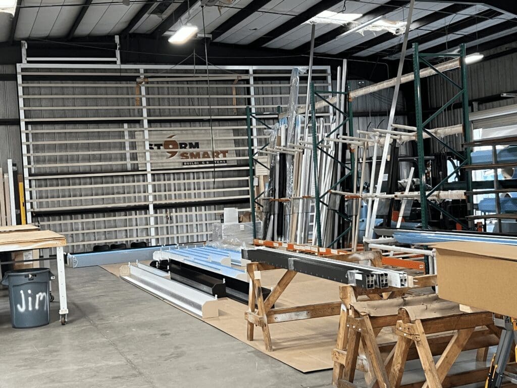 A cluttered workshop with various construction materials like metal frames, ladders, and beams designed for impact resistance. There are sawhorses, a wooden pallet, and a sign that says "STORM SMART. in either North or South Carolina