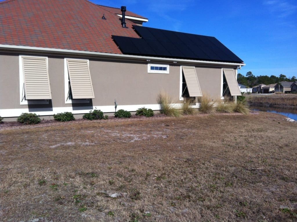 A single-story house with beige exterior walls and hurricane shutters. The roof features solar panels. There's a small pond at the back and grassy, patchy land in the foreground under a clear blue sky. In either north or south carolina - american hurricane shutters A single-story house with beige exterior walls and hurricane shutters. The roof features solar panels. There's a small pond at the back and grassy, patchy land in the foreground under a clear blue sky. in either North or South Carolina