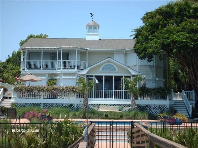 Two-story coastal house with light green siding, white trim, and multiple gables, featuring a covered porch, palm trees, and a well-kept garden, illustrating a serene environment suitable for hurricane protection solutions.