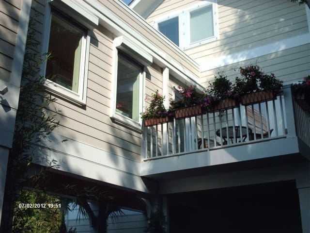 Two-story coastal house with light-colored siding, featuring windows with shutters, a balcony adorned with flowers, and a covered porch, illustrating hurricane protection solutions.