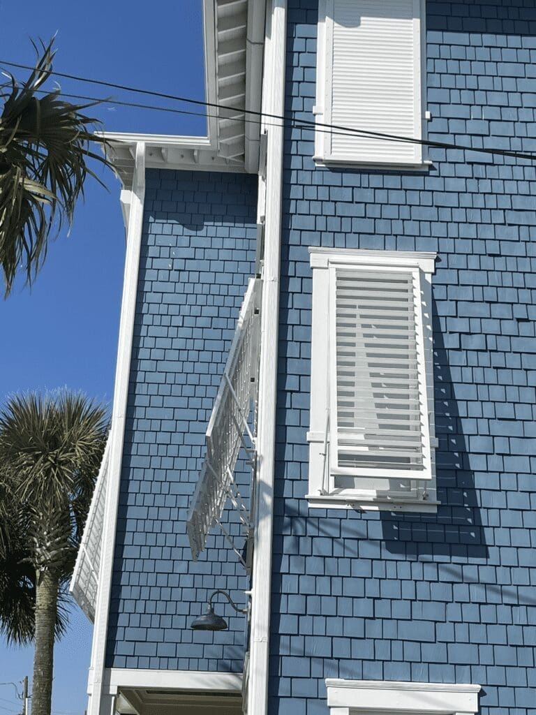 - american hurricane shutters House with blue shingle siding featuring white Bahama shutters and adjustable louvers, surrounded by palm trees, emphasizing coastal design and hurricane protection.