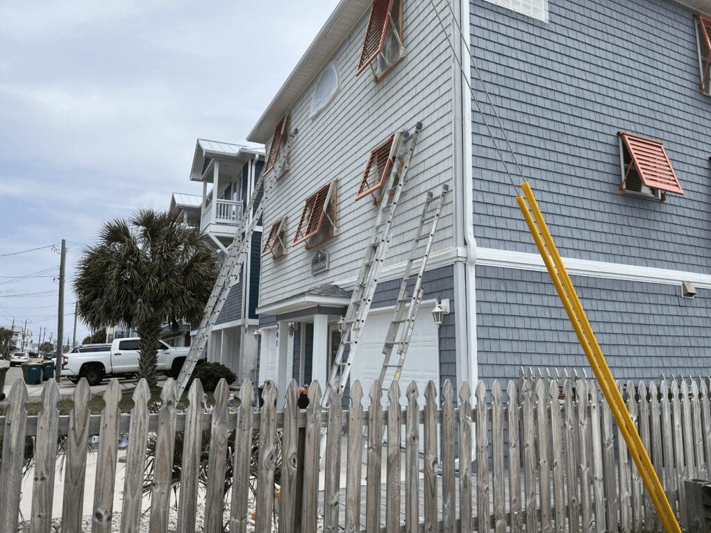Pink articulating bahama shutters - american hurricane shutters Ladders leaning against a multi-story blue building with pink Articulating Bahama Shutters, viewed from behind a weathered wooden picket fence. A palm tree and clear sky are visible. in either North or South Carolina