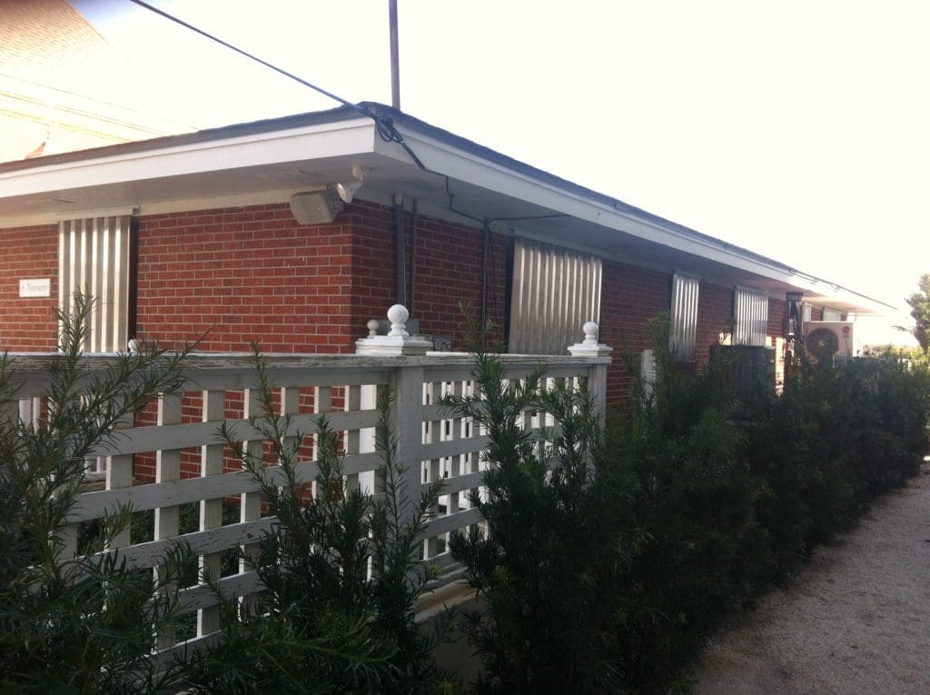 A row of single-story red brick motel units with white fences and green bushes in front, captured on a sunny day in Carolina. in either North or South Carolina