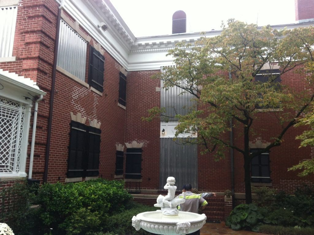 Historic courtyard building with hurricane shutters, wilmington - american hurricane shutters Hurricane panels installed on a brick home, showcasing aluminum protection against storm damage, with a garden fountain and greenery in the foreground.