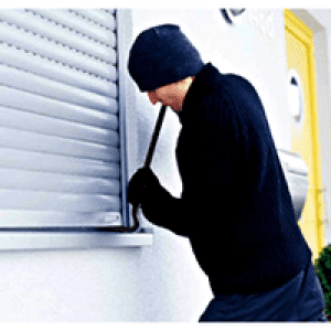 A person wearing a black beanie and dark clothing is using a tool to break into a house through a window with hurricane shutters. in either North or South Carolina