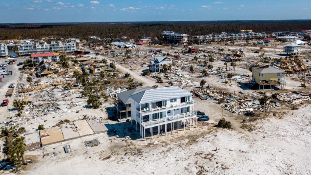 Aerial view of a coastal Carolina area showing extensive hurricane impact. Many homes are destroyed or severely damaged, with debris scattered around. One house remains intact amid the devastation. in either North or South Carolina