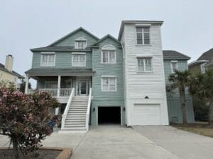 A two-story coastal house with light green and white siding. It features multiple gables, a covered front porch with white railings, and a single-car garage. There are steps leading to the front door and a palm tree and blooming bush in the front yard. Hurricane Shutters in Jacksonville