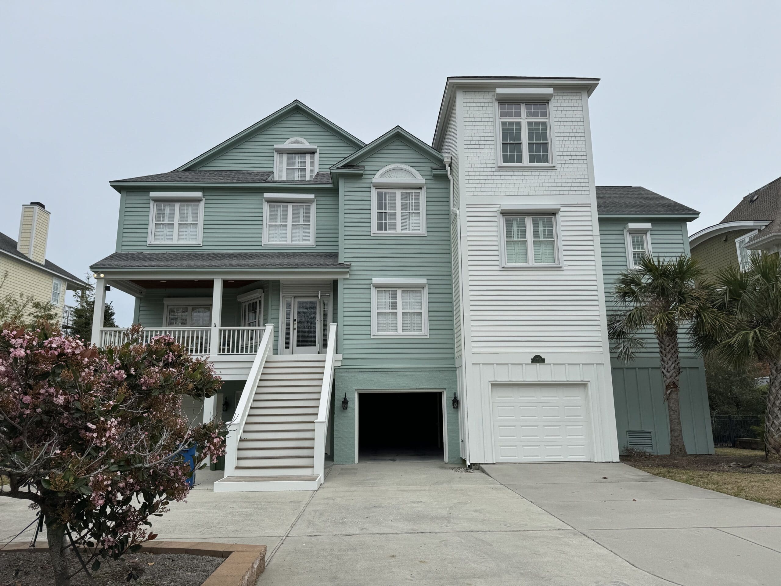 A two-story coastal house with light green and white siding. It features multiple gables, a covered front porch with white railings, and a single-car garage. There are steps leading to the front door and a palm tree and blooming bush in the front yard. in either North or South Carolina