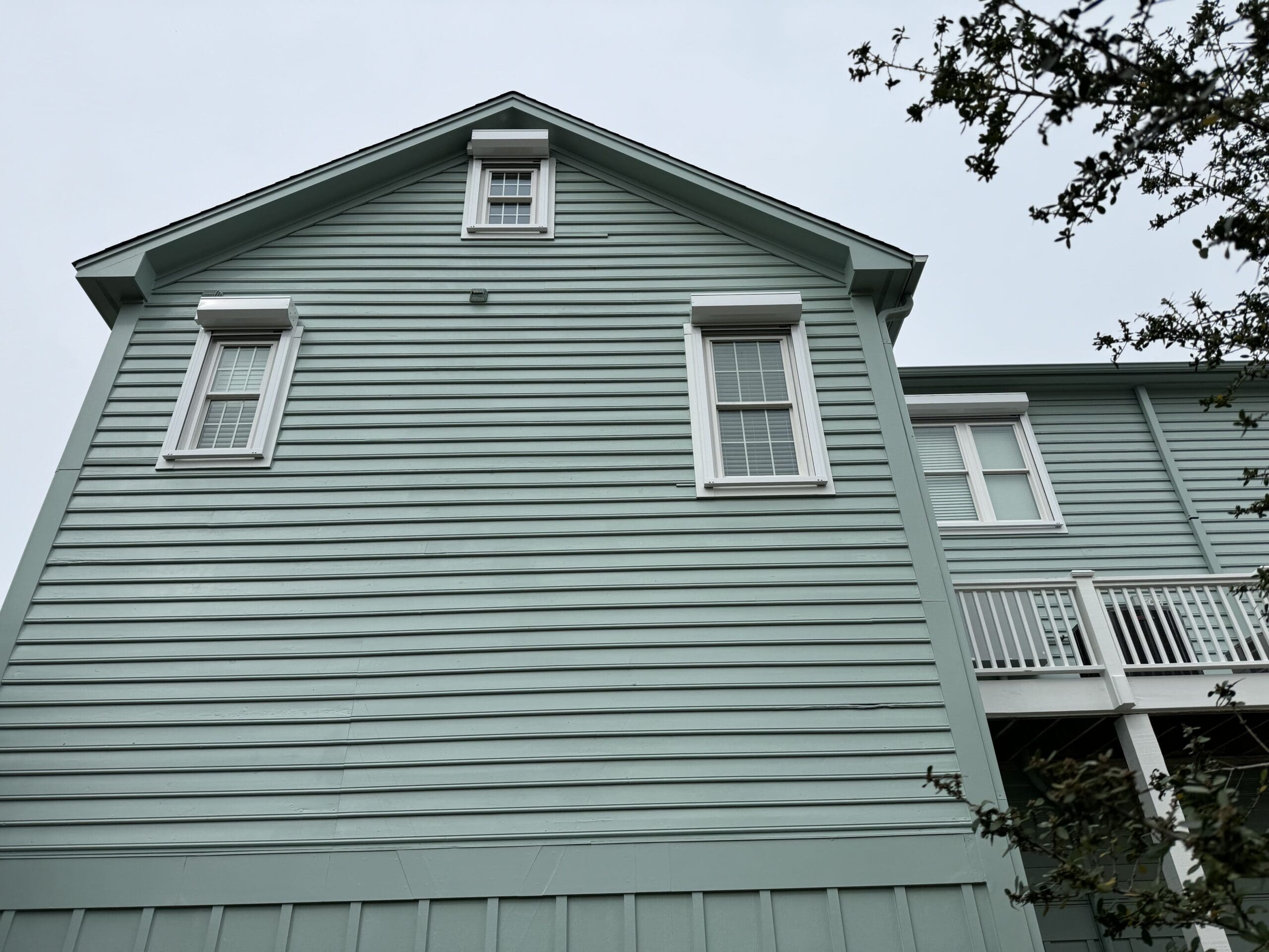 A two-story house features light blue-green siding with white trim. The photo captures an angled upward view of the structure, highlighting two windows with shutters and a small gable window in the upper section. A leafless tree branch is visible to the right. in either North or South Carolina