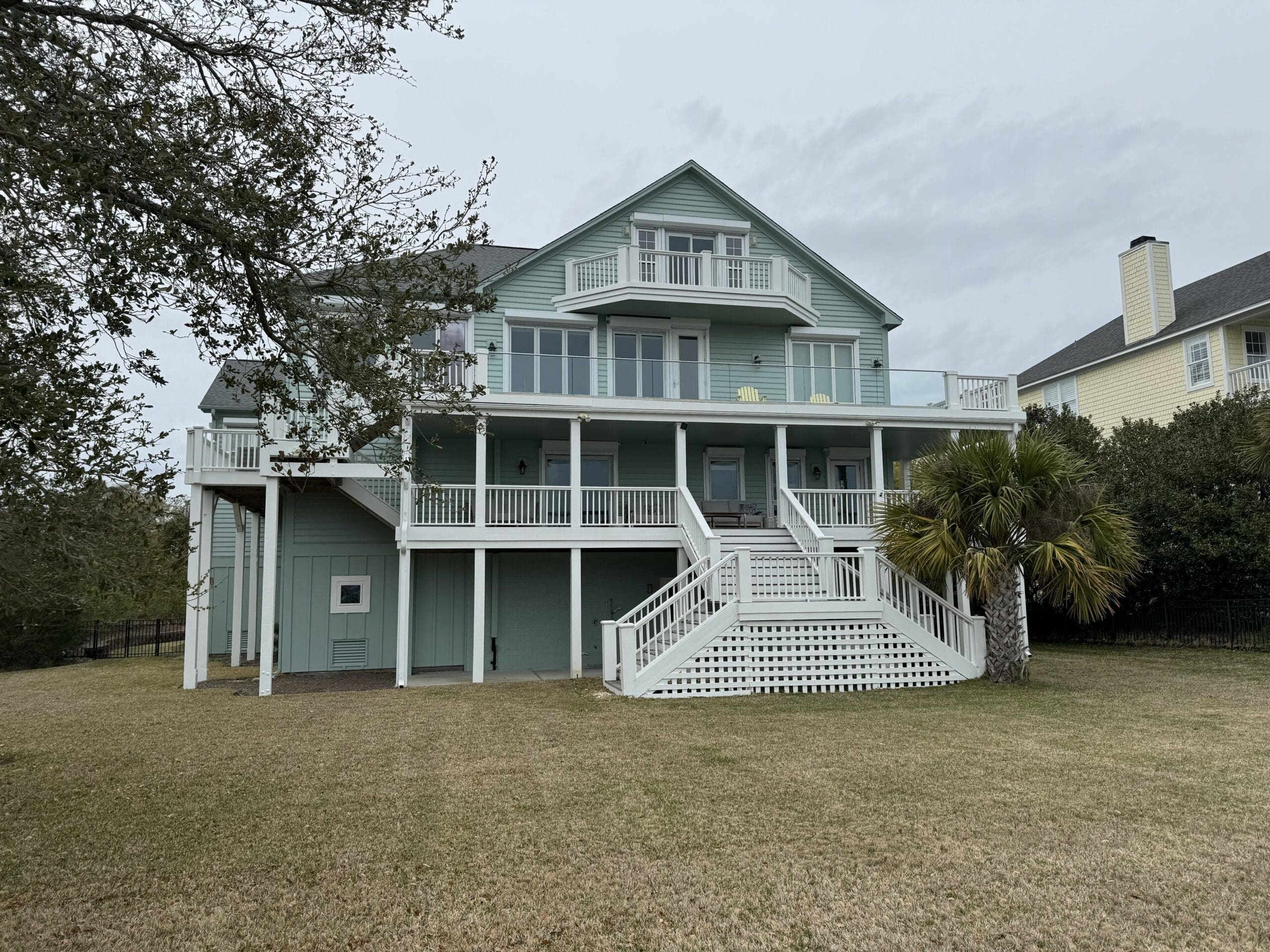 A two-story green house with a wide staircase leading to a large deck and multiple balconies. The house has a sloped roof and is surrounded by a well-kept grassy yard with trees, including a palm tree on the right. The sky is overcast. in either North or South Carolina