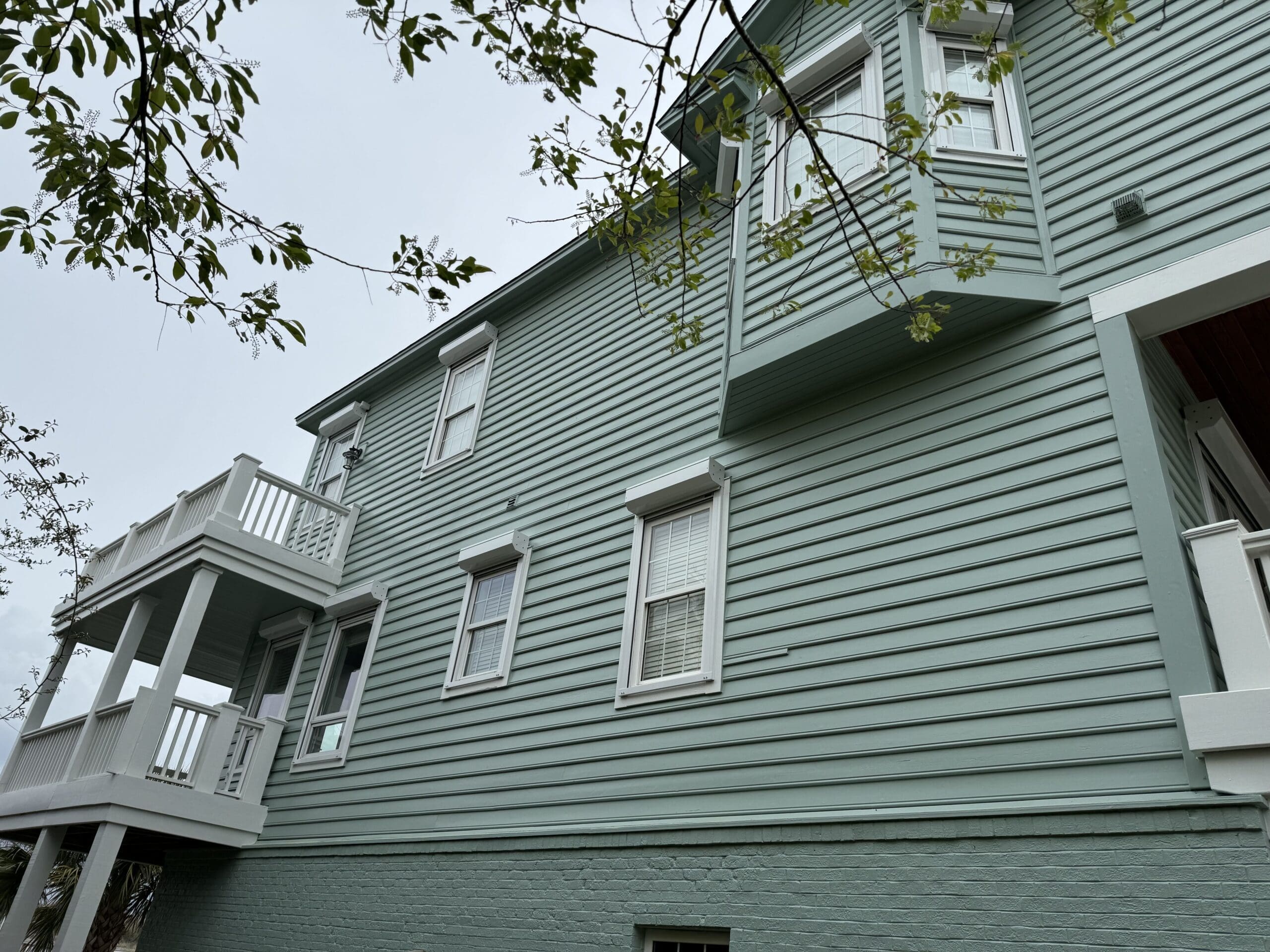 A light green, two-story house with white-trimmed windows and a protruding bay window. A balcony extends from the left side of the house, supported by white columns. A tree branch with green leaves hangs in the foreground. in either North or South Carolina