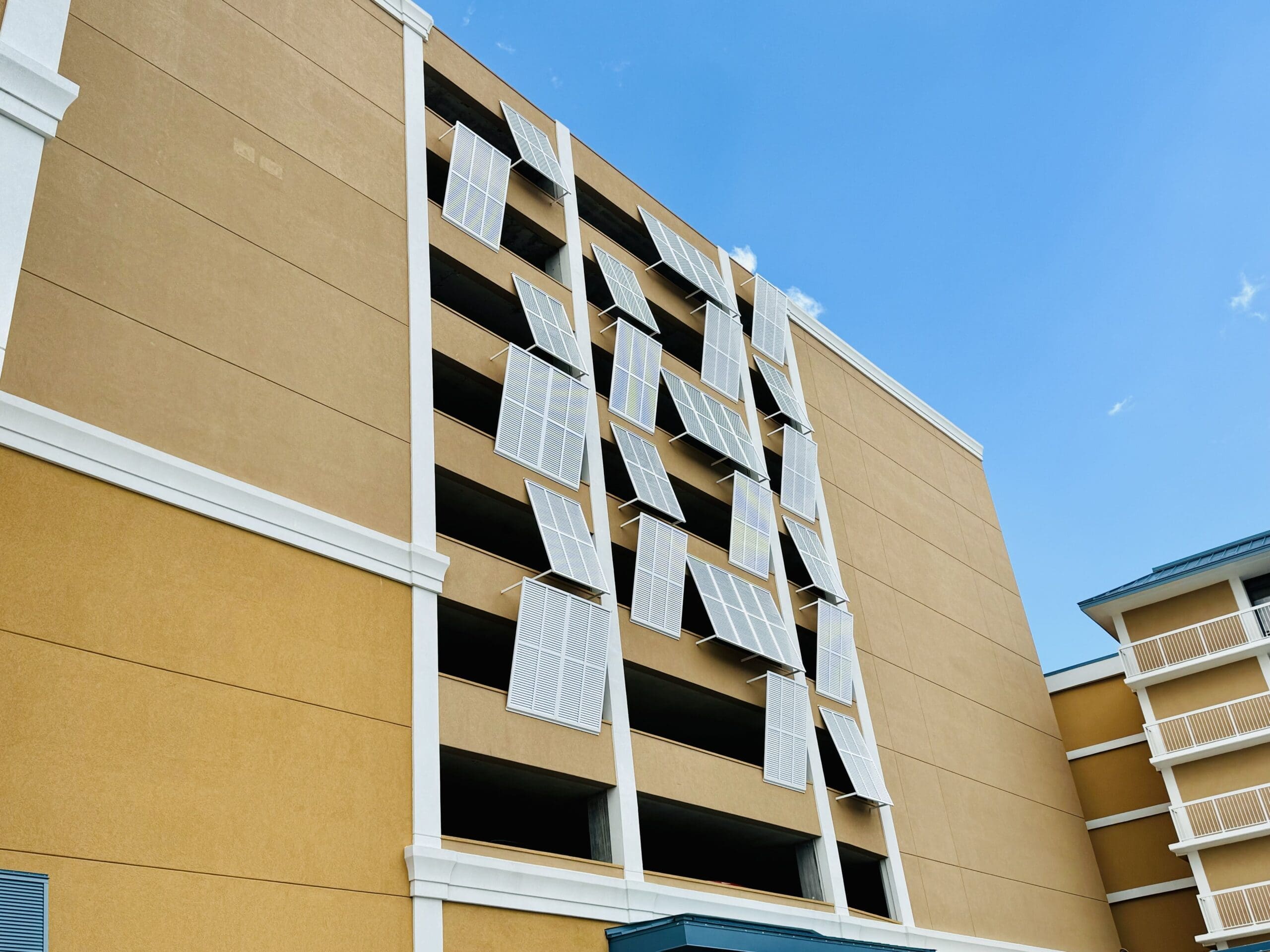 A multi-story building with large window shutters hanging loosely, appearing damaged. The building has beige walls with white accents along the edges. The sky is clear and blue in the background. in either North or South Carolina