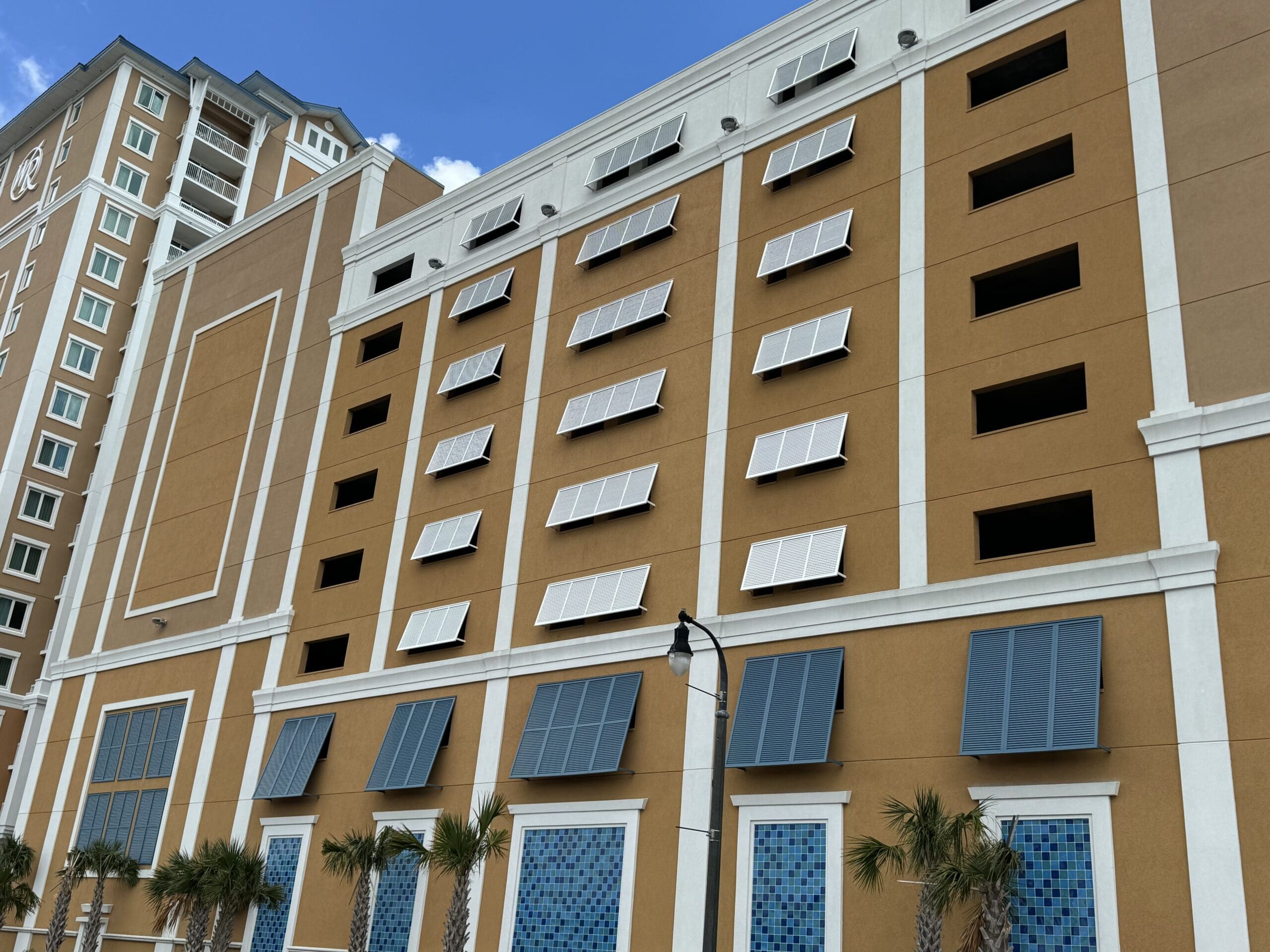 A multi-story building with light brown and white exterior walls featuring rows of panels and large windows. Palm trees line the front, and a street lamp stands in front of the building. A tall section of the building extends to the left. The sky is clear and blue. in either North or South Carolina