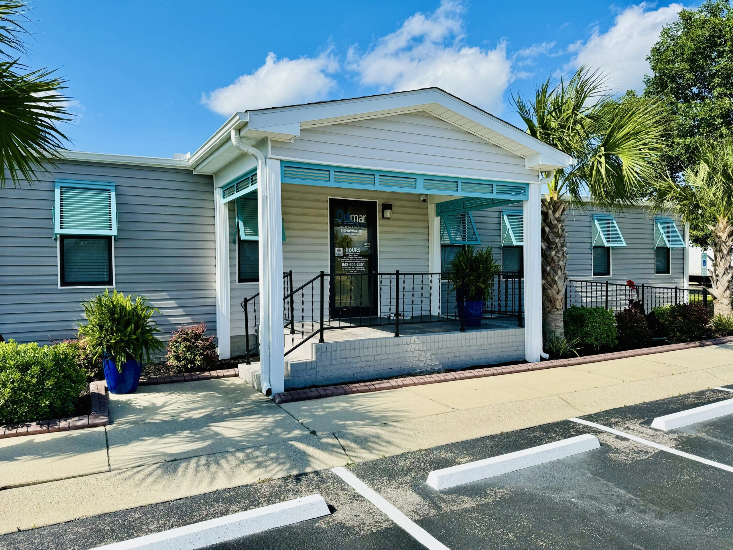 A single-story, modern building with light gray siding and white trim, featuring a front porch with steps and black railing. Palm trees and blue potted plants decorate the exterior. A parking lot with marked spaces is in the foreground, and the sky is clear and sunny. in either North or South Carolina