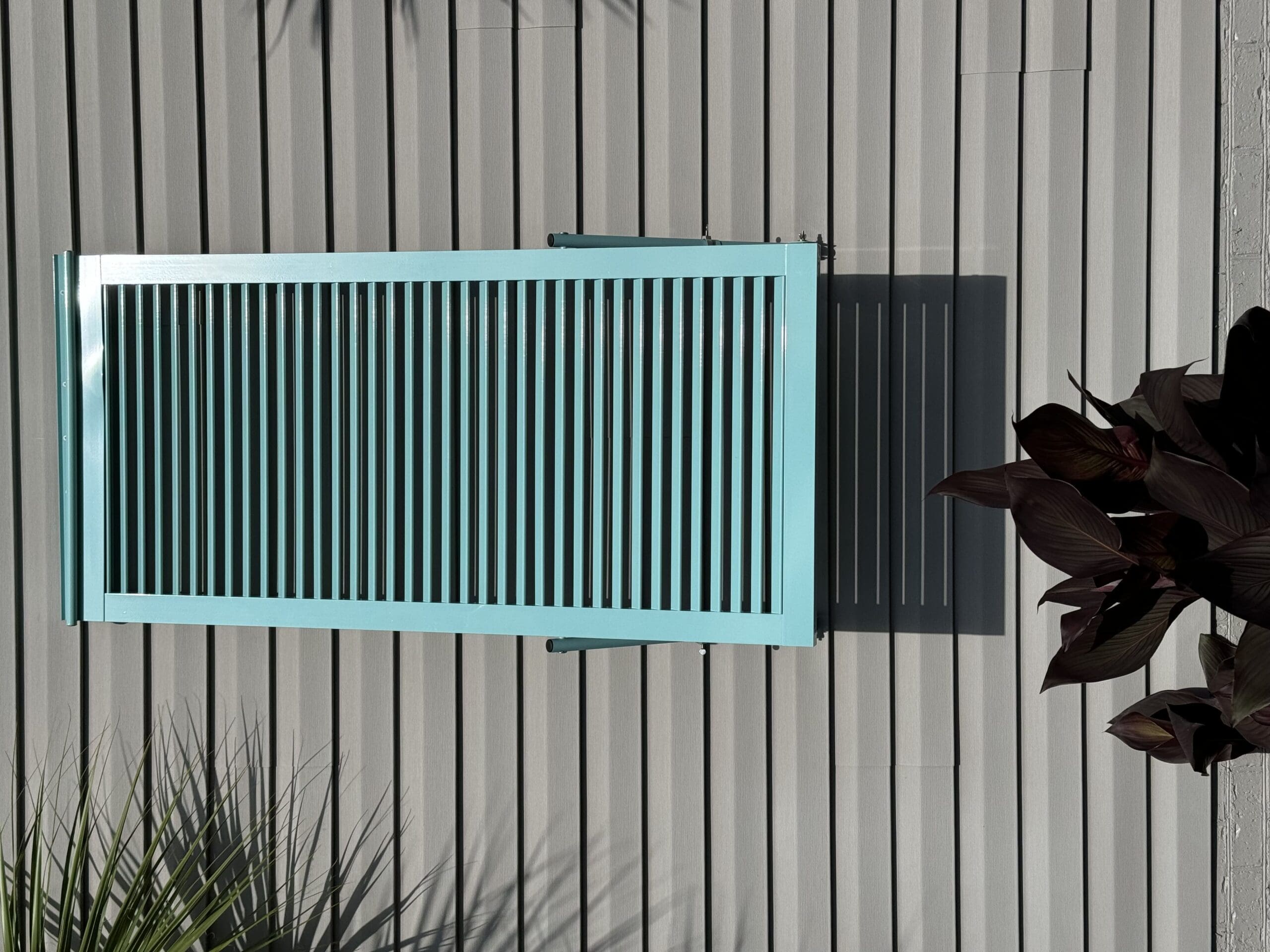 A closed turquoise shutter attached to a light gray, horizontally paneled exterior wall. A small potted plant with dark leaves sits below, and a palm tree is partially visible on the left side, casting shadows on the wall. in either North or South Carolina