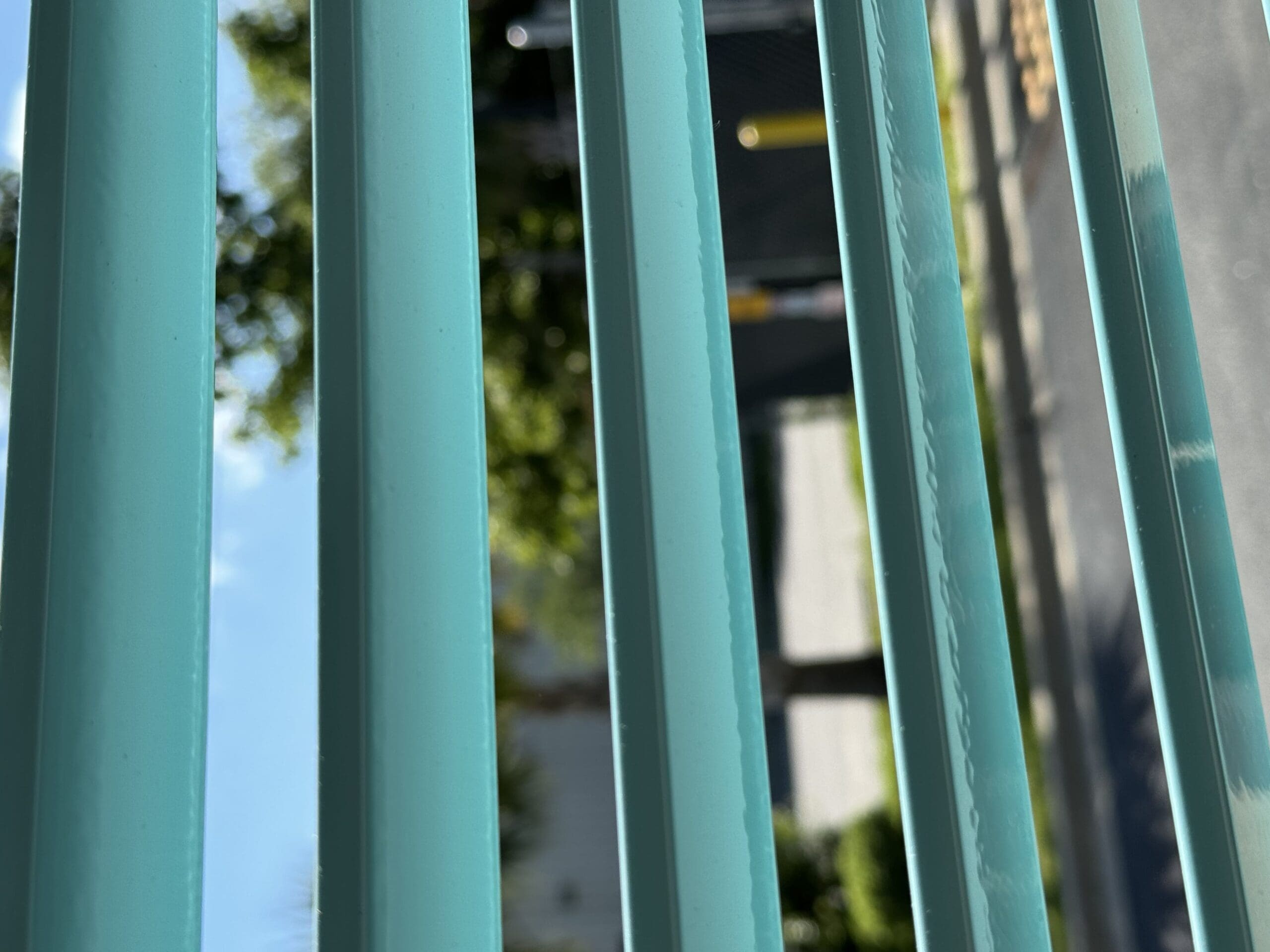 Close-up view through a set of turquoise window blinds, showing an outdoor scene. The background reveals a blurry image of a street, a few trees, and other structures bathed in sunlight. The focus is on the blinds, with the outdoor elements providing a soft backdrop. in either North or South Carolina