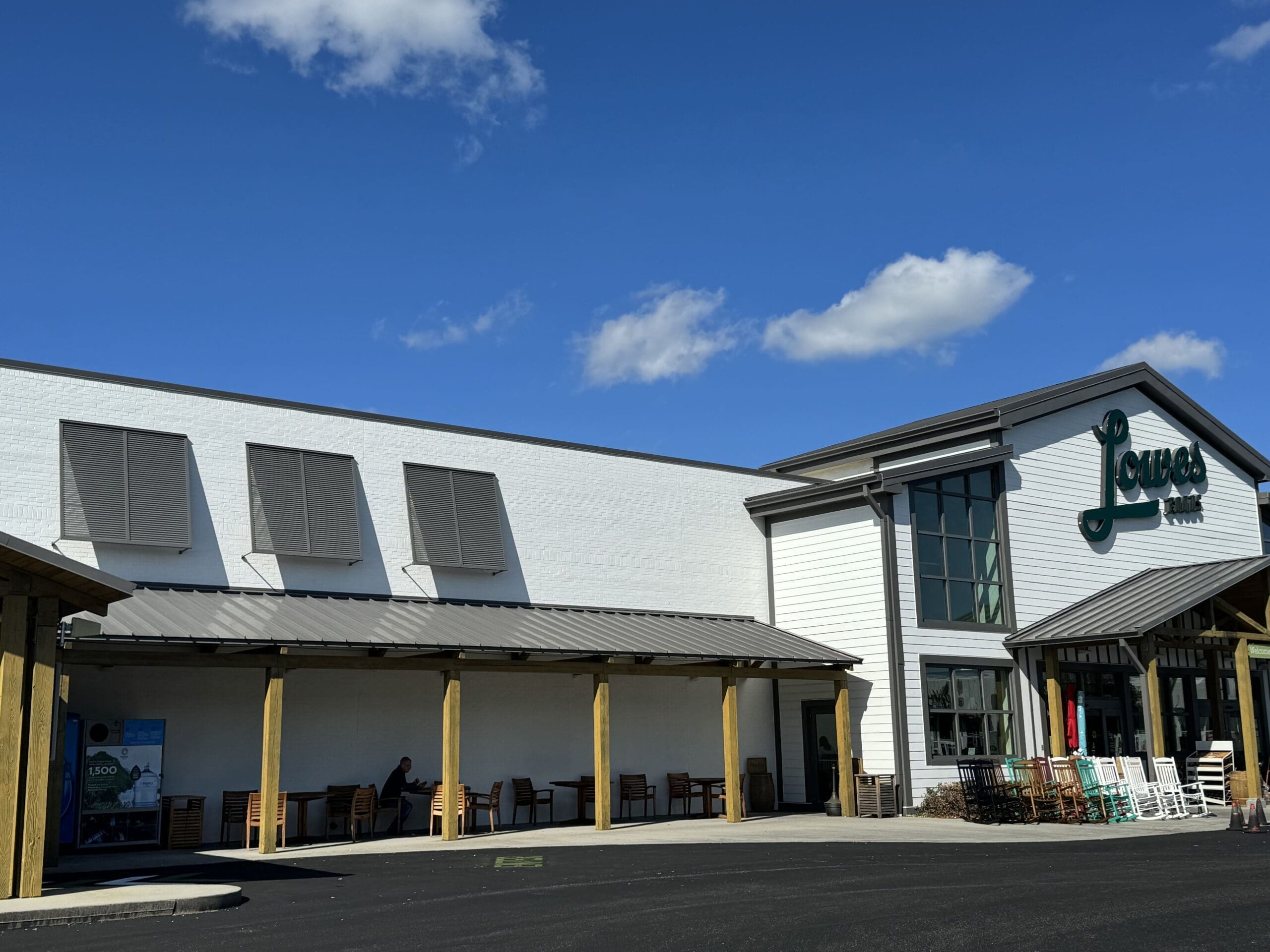 A white building with a sign reading "Lowe's" on the front. The facade includes large windows and a wooden overhang covering an outdoor seating area. The sky is clear and blue with a few clouds. A single person sits at a table in the shaded seating area. in either North or South Carolina