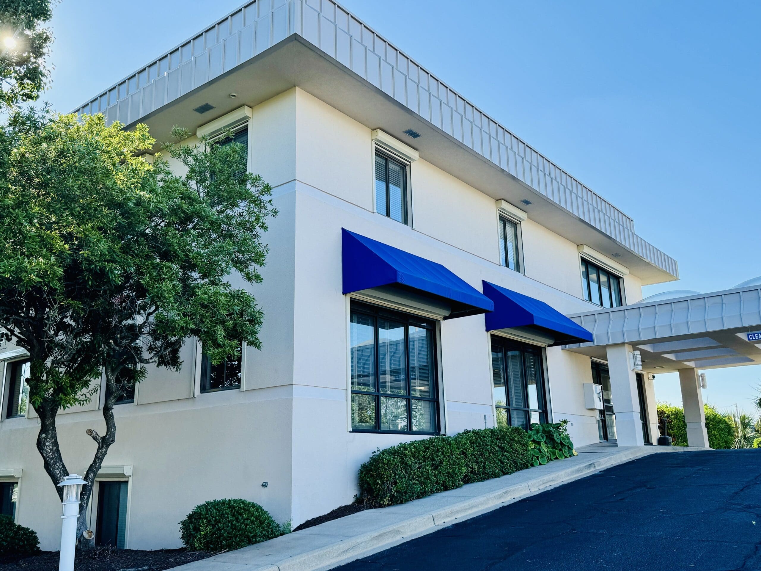 A two-story white building with blue awnings over the windows, surrounded by greenery. The building sits on a slight incline with a paved driveway leading to a covered entrance on the right side. The sky above is clear and blue. in either North or South Carolina