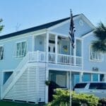 A light blue, two-story house is shown on a sunny day. The house features a front porch with stairs leading up to it, white trim, and shuttered windows. An American flag flies in the yard next to a palm tree, and a white SUV is parked in the driveway. in either North or South Carolina