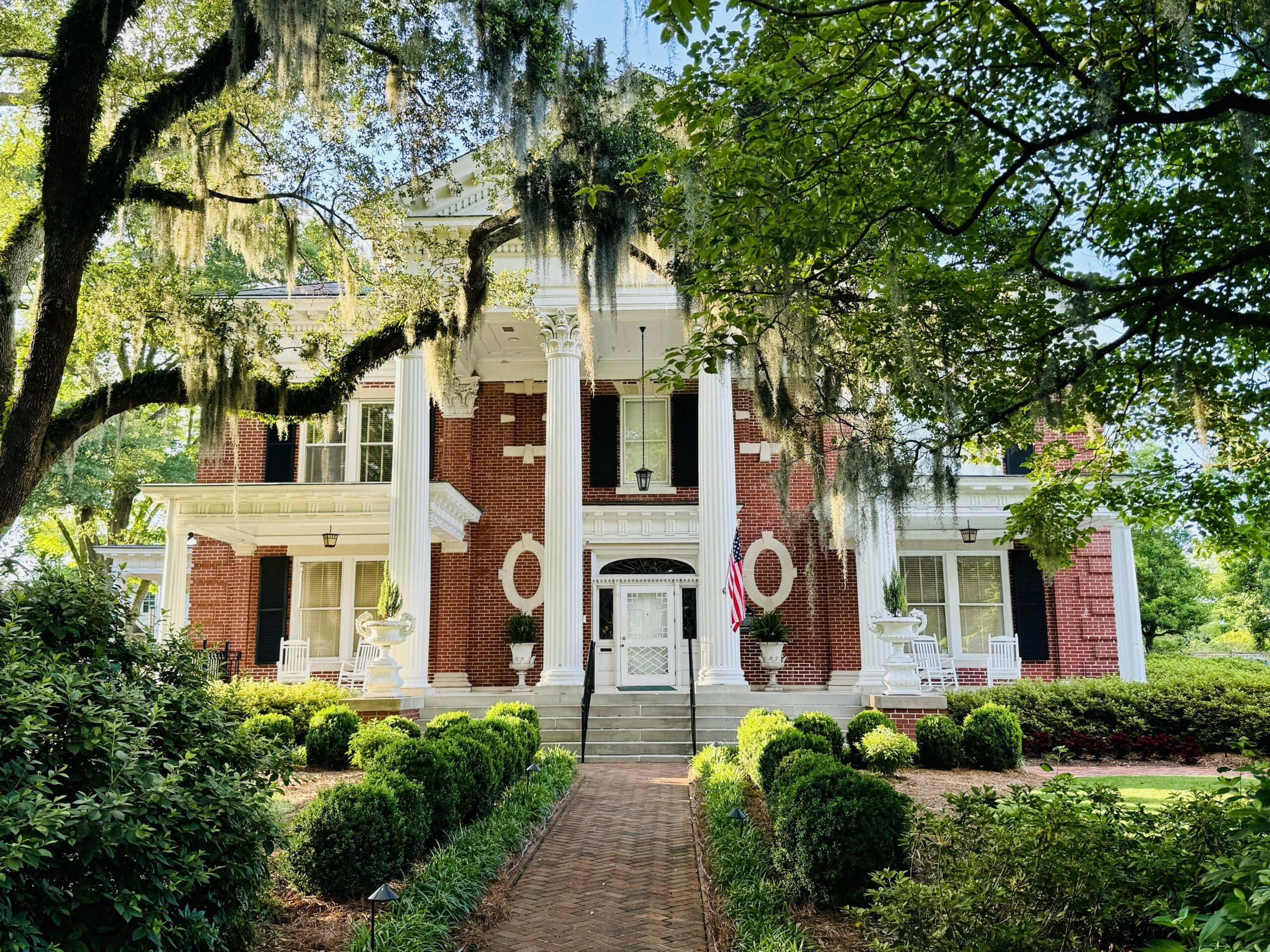 A grand, two-story brick mansion with white columns and ornate detailing stands amidst lush greenery and trees. Moss hangs from branches, creating a historic Southern ambiance. A brick path leads to the entrance, flanked by manicured bushes. in either North or South Carolina