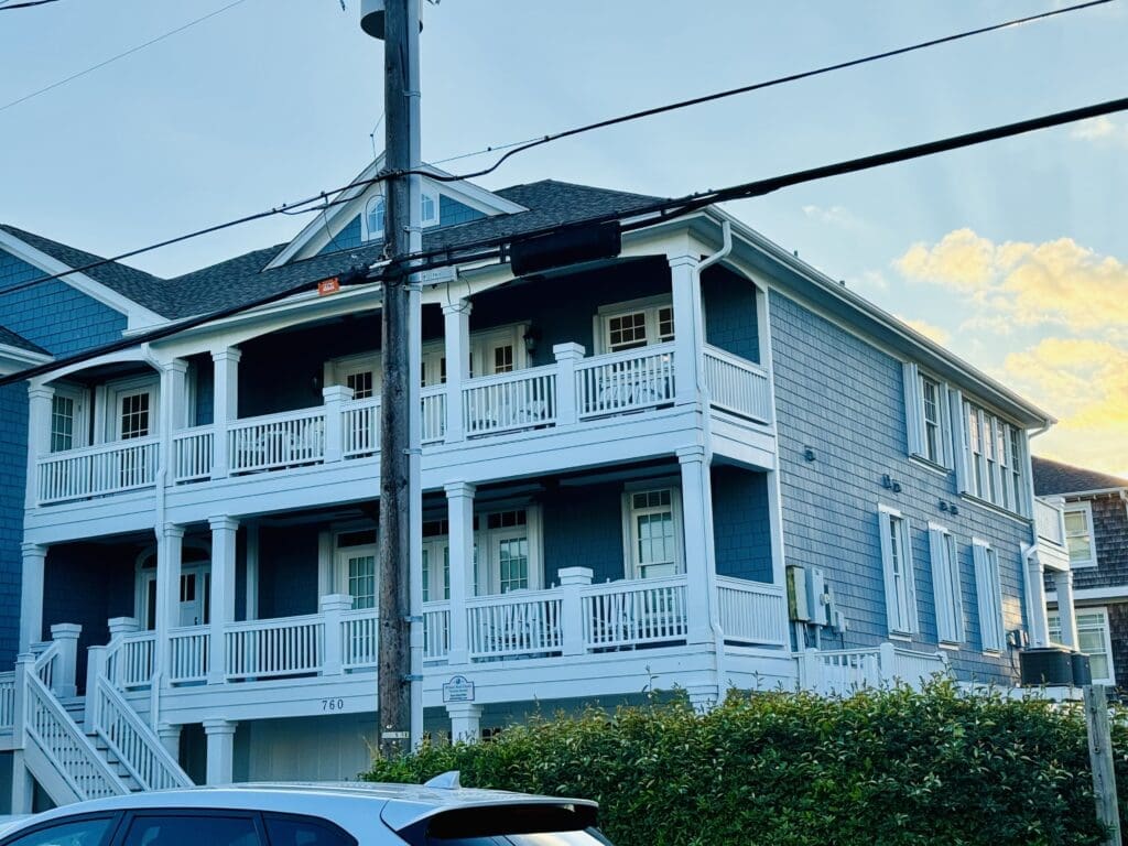 Two-story blue house with white trim and multiple wrap-around balconies, set against a clear sky. Steps lead up to the main entrance on the left. A car is parked in the foreground, and power lines cross the middle of the scene. Bushes line the front yard. Shows accordion hurricane shutters in Morehead City.