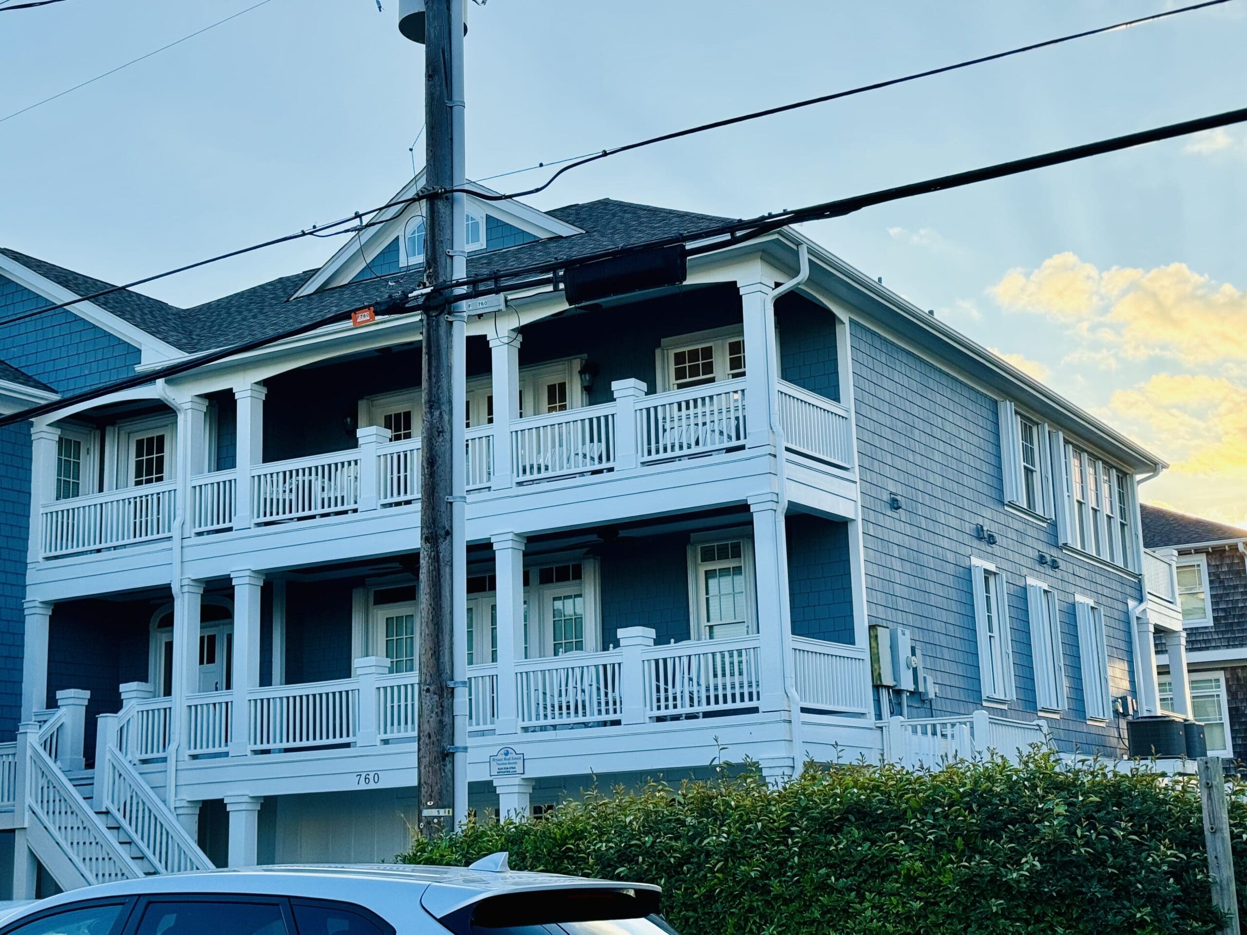 Two-story blue house with white trim and multiple wrap-around balconies, set against a clear sky. Steps lead up to the main entrance on the left. A car is parked in the foreground, and power lines cross the middle of the scene. Bushes line the front yard. in either North or South Carolina