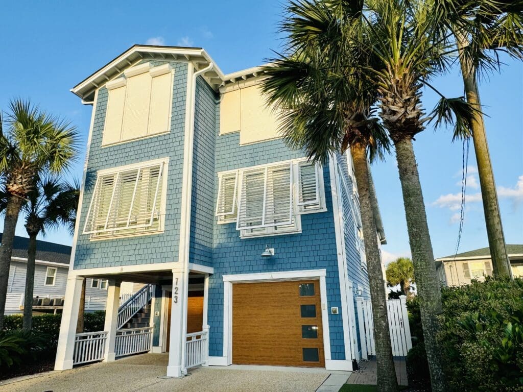 A two-story blue beach house with white trim, featuring large windows with Bahama shutters. There is a wooden garage door on the lower level. Palm trees surround the property, and the sky is clear with a few clouds visible. The house number "123" is displayed near the entrance. in either North or South Carolina