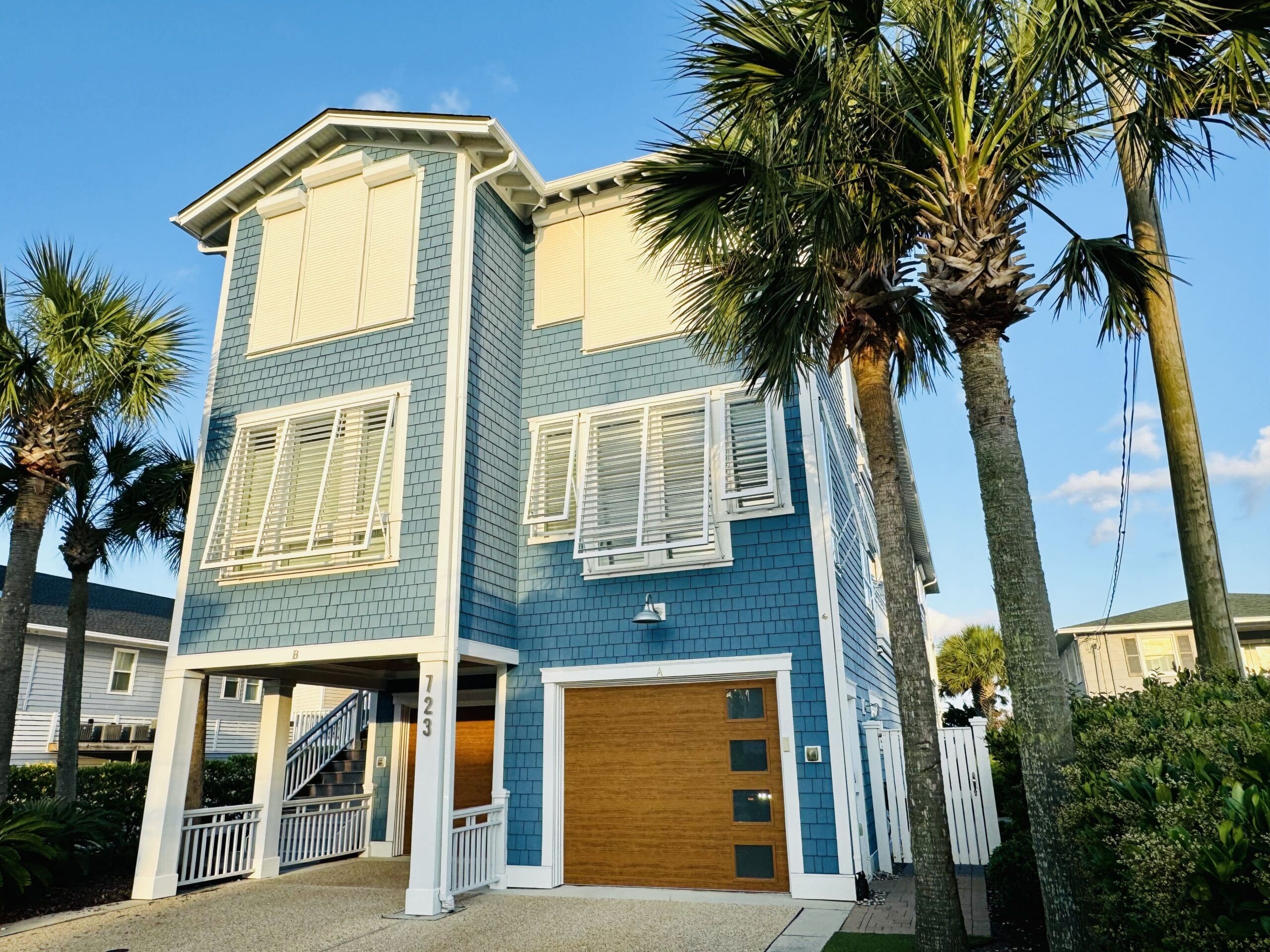 A two-story blue beach house with white trim, featuring large windows with shutters. There is a wooden garage door on the lower level. Palm trees surround the property, and the sky is clear with a few clouds visible. The house number "123" is displayed near the entrance. in either North or South Carolina