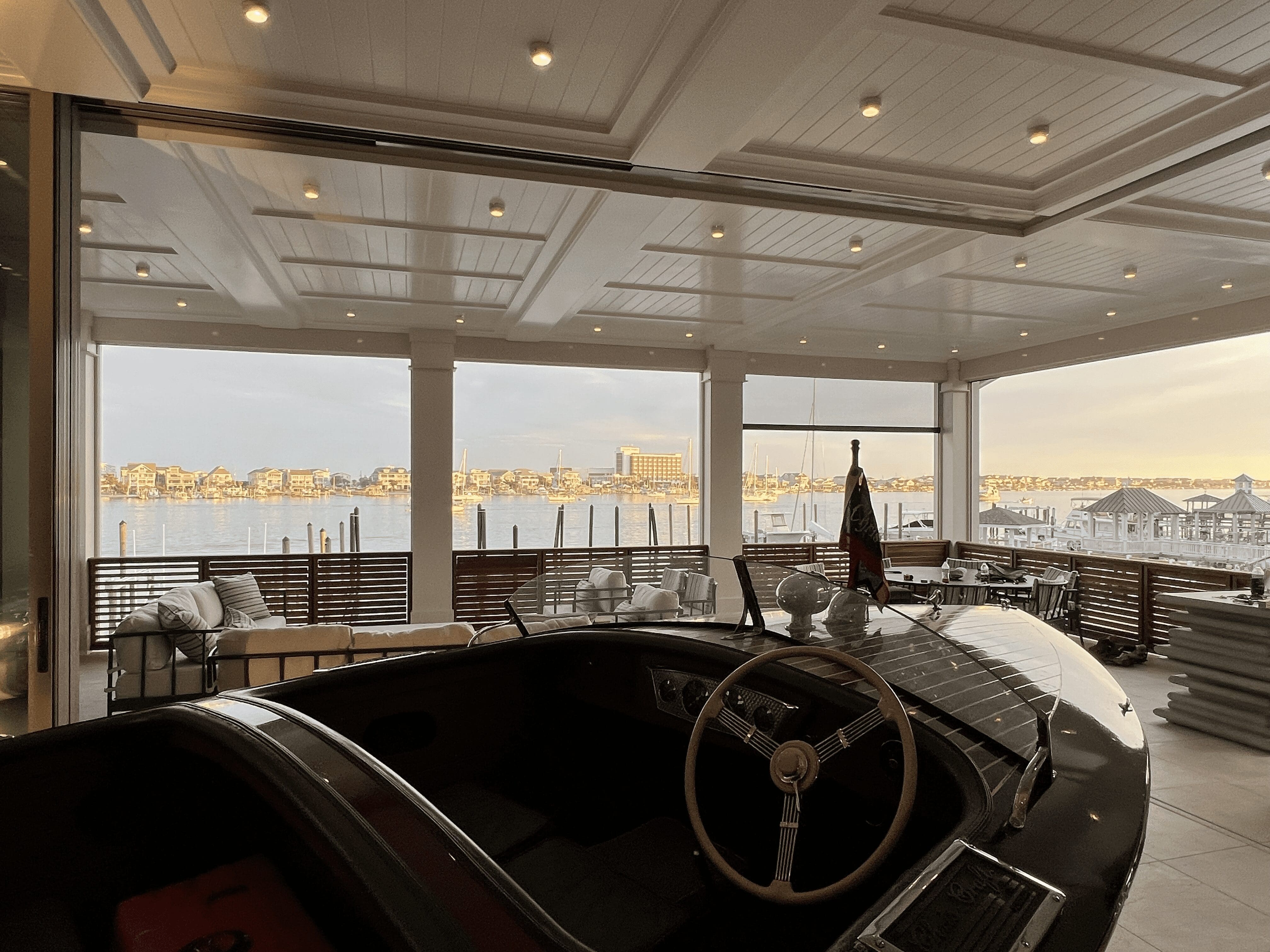 A luxurious, covered veranda overlooks a scenic marina with numerous docked boats. The area is well-lit with recessed lighting. In the foreground, part of a classic car's interior with a wooden steering wheel is visible, adding a touch of vintage elegance. in either North or South Carolina