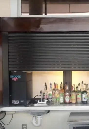 A small bar area with a half-closed metal shutter reminiscent of hurricane shutters hovers above a counter filled with various liquor bottles. A "Budweiser" branded sign is partially visible on the left. A sink with a faucet is tucked underneath the counter. in either North or South Carolina