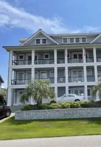 A three-story gray building with white trim and hurricane shutters, featuring balconies adorned with American flags. A white car is parked in front, and the property boasts palm trees and a well-kept lawn. The sky is clear and blue. in either North or South Carolina