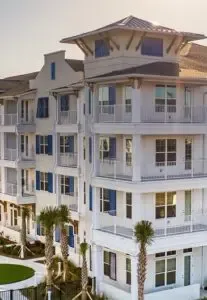 White multi-story beachside building with blue shutters and balconies, thoughtfully equipped with hurricane shutters, framed by palm trees. The architecture showcases a mix of traditional and modern elements under a sunny sky. in either North or South Carolina