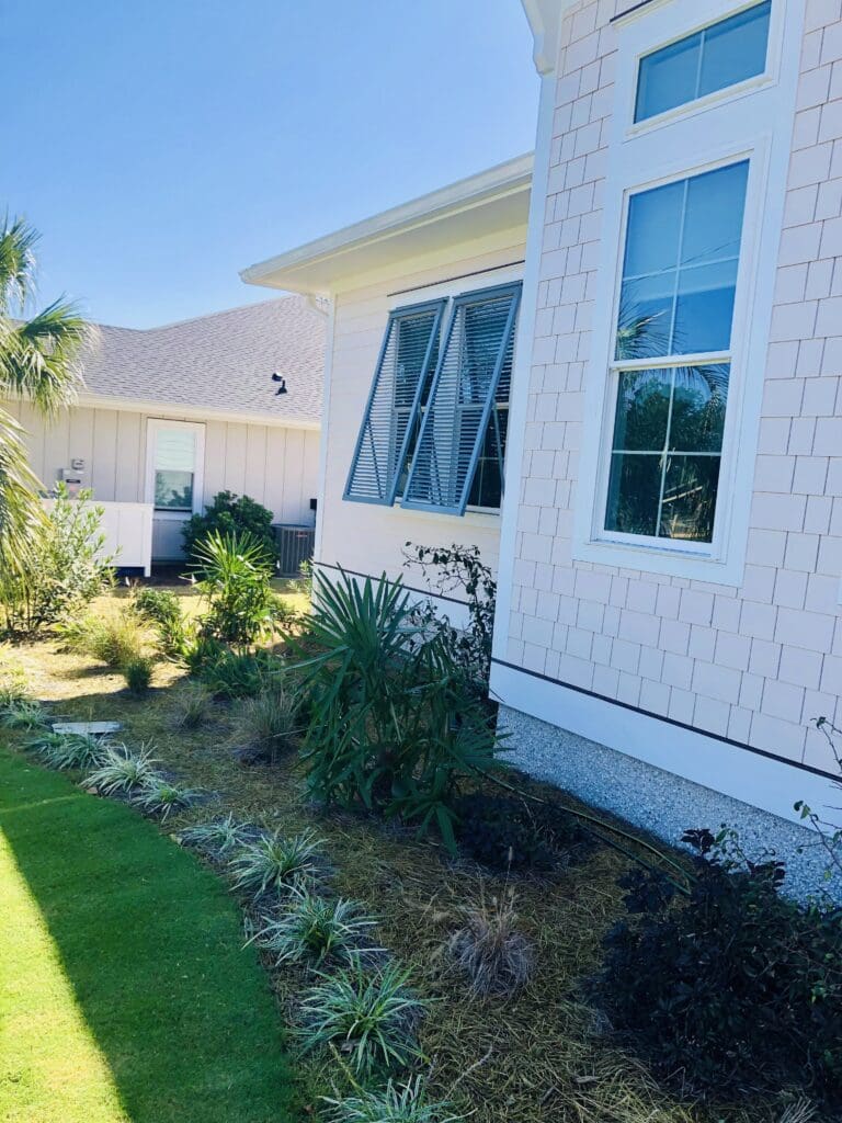 A modern house exterior in NC showcases large windows with beautiful bahama shutters and a neatly landscaped garden with various plants and shrubs. The light-colored siding complements the hurricane shutters, while the clear blue sky casts shadows on the lawn, reminiscent of serene days at Topsail Beach, North Carolina.