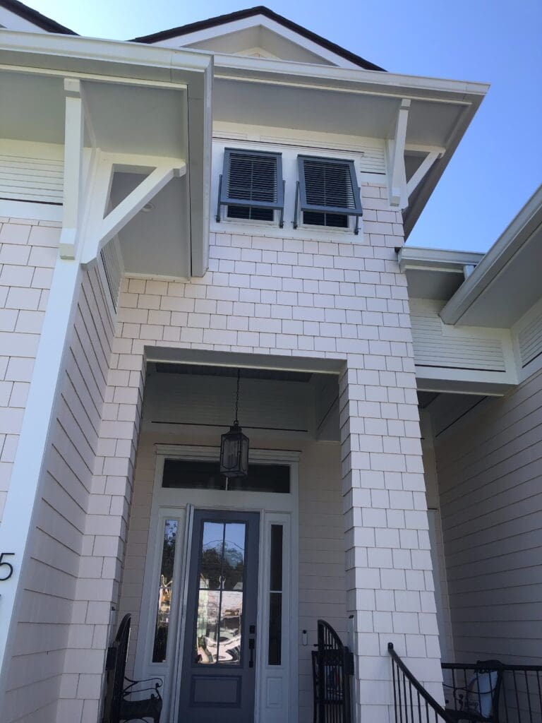 A two-story house with light-colored siding and dark shutters, including stylish Bahama shutters. It features a front entrance with a glass-paneled door, a hanging lantern, and black wrought iron railings. The roof has gables, and the sky is clear and blue. in either North or South Carolina A two-story house with light-colored siding and dark shutters, including stylish Bahama shutters. It features a front entrance with a glass-paneled door, a hanging lantern, and black wrought iron railings. The roof has gables, and the sky is clear and blue. in either North or South Carolina