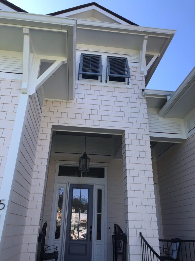 A two-story house with light-colored siding and dark shutters, including stylish Bahama shutters. It features a front entrance with a glass-paneled door, a hanging lantern, and black wrought iron railings. The roof has gables, and the sky is clear and blue. in either North or South Carolina