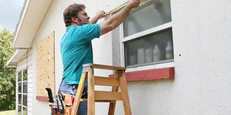 A person in a teal shirt stands on a wooden ladder, measuring a window for residential storm shutters on a house's exterior. Their tool belt is filled with various tools. The window is partially shaded, and another nearby is boarded up. In the background, trees sway gently near Wrightsville Beach. in either North or South Carolina