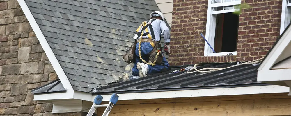 A construction worker, outfitted in safety gear, is kneeling on a house roof at Wrightsville Beach, expertly using tools. The gray-shingled roof with its metal section overlooks a window and brick facade, while a ladder leans against the house—perhaps an investment in the making. in either North or South Carolina