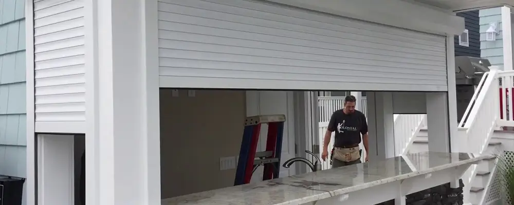 A garage space in South Carolina is being transformed into a bar with a granite counter. A partially open white roller shutter exposes a man working inside, perhaps planning to add Lexan Hurricane Panels. White siding and stairs of this coastal homeowner's dream are visible in the background. in either North or South Carolina