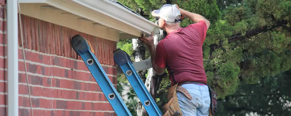 A man in a red shirt, jeans, and cap is meticulously maintaining the gutter of a brick house. He stands on a blue ladder, tools snugly in his belt, with lush trees adding charm reminiscent of Myrtle Beach around him. in either North or South Carolina