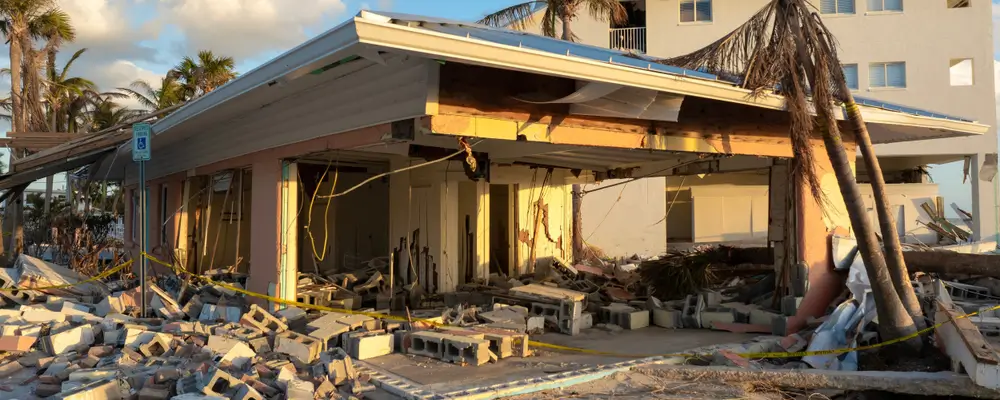 A building with a collapsed roof and walls, surrounded by debris and caution tape, highlights the challenges Wilmington homeowners face. Palm trees stand nearby, suggesting recent damage from a natural disaster. The clear sky offers little solace as electric rolldown shutters become top trends for protection. in either North or South Carolina