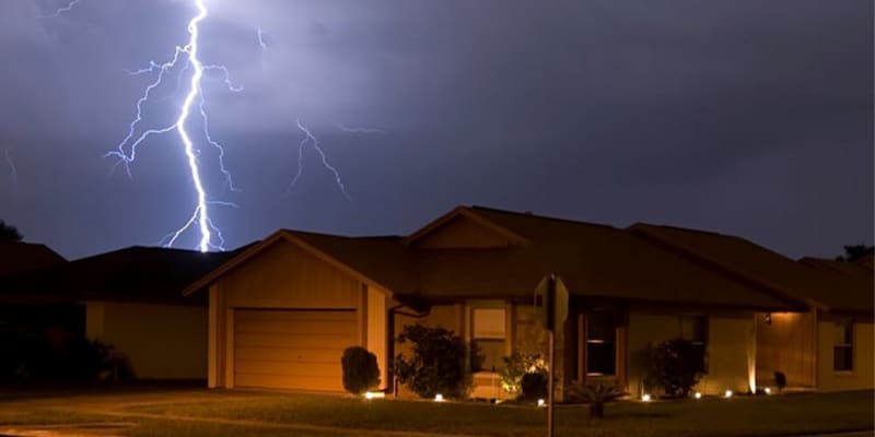 A single-story house in Wilmington is illuminated under a dark, stormy sky by a bright lightning bolt striking in the background. The house has a garage and a small garden, with exterior lights casting a warm glow, showcasing the residents' dedication to storm protection. in either North or South Carolina