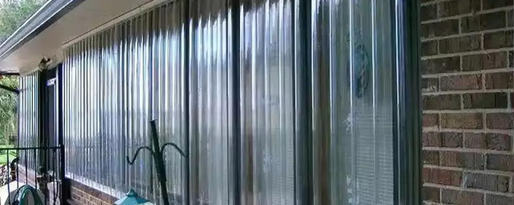 Wilmington home with clear corrugated hurricane shutters - american hurricane shutters Aluminum storm shutters and Lexan hurricane panels safeguard a long set of windows on this brick exterior, offering coastal homeowners in South Carolina peace of mind. A porch with metal railings and outdoor furniture nestles comfortably under the roof's overhang, providing a shaded retreat. in either North or South Carolina