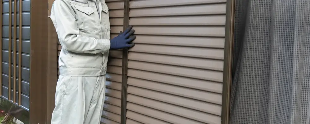 Worker installs slatted hurricane shutters, topsail - american hurricane shutters A person in a light gray work uniform and dark gloves stands beside a set of brown horizontal slatted blinds, reminiscent of residential storm shutters. Behind the blinds, a mesh curtain is partially visible. The setting seems to be outdoors or in a garden area near Wrightsville Beach. in either North or South Carolina