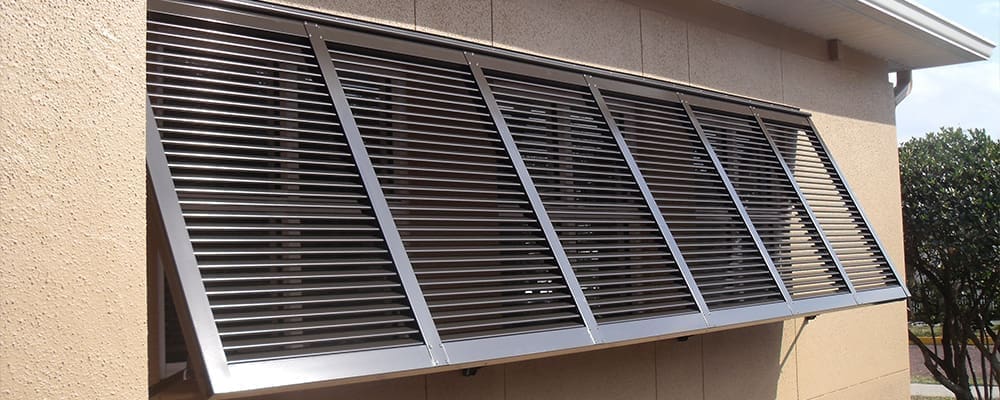 A sleek aluminum louvered sunshade, reminiscent of Bahama Shutters, is mounted above a window on a beige building. The slanted design allows airflow while providing shade and adding style. Part of a tree is visible to the right of the building, enhancing home protection naturally. in either North or South Carolina