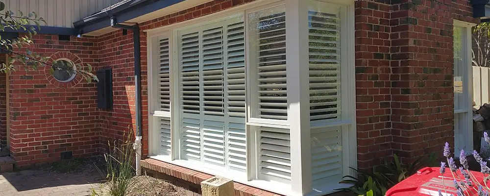 A red-brick house in Myrtle Beach features a large window with white plantation shutters. Nearby, a small circular window and some potted plants are visible. A red recycling bin is in the foreground, hinting at the charm of maintaining manual accordion shutters nearby. in either North or South Carolina