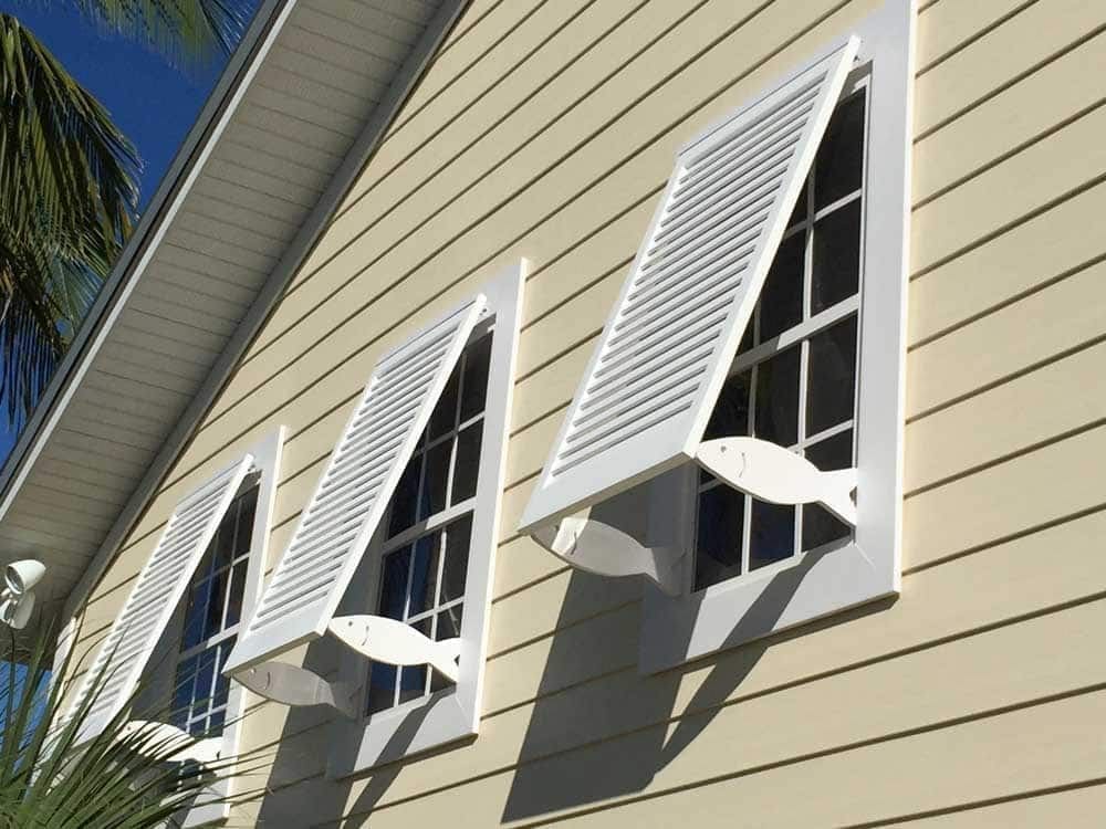 Fish-cutout Bahama hurricane shutters on a yellow coastal home.