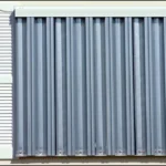 A window in New Bern covered with energy-efficient storm shutters, flanked by closed white louvered shutters, on a beige paneled exterior wall—showcasing custom hurricane solutions for ultimate protection and style. in either North or South Carolina