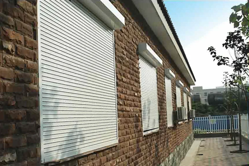 A brick building with white rolldown storm shutters closed over its windows; trees and a fence line the paved path beside it. in either North or South Carolina