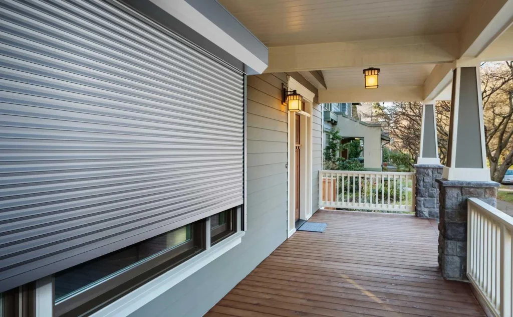 A porch with wooden flooring features a closed silver rolldown storm shutter over a window, gray siding, white railings, stone pillars, and two hanging lantern-style lights near the entrance. in either North or South Carolina