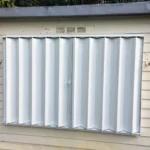 A white accordion-style metal hurricane shutter is closed over a window on the exterior of a beige house with horizontal siding. in either North or South Carolina